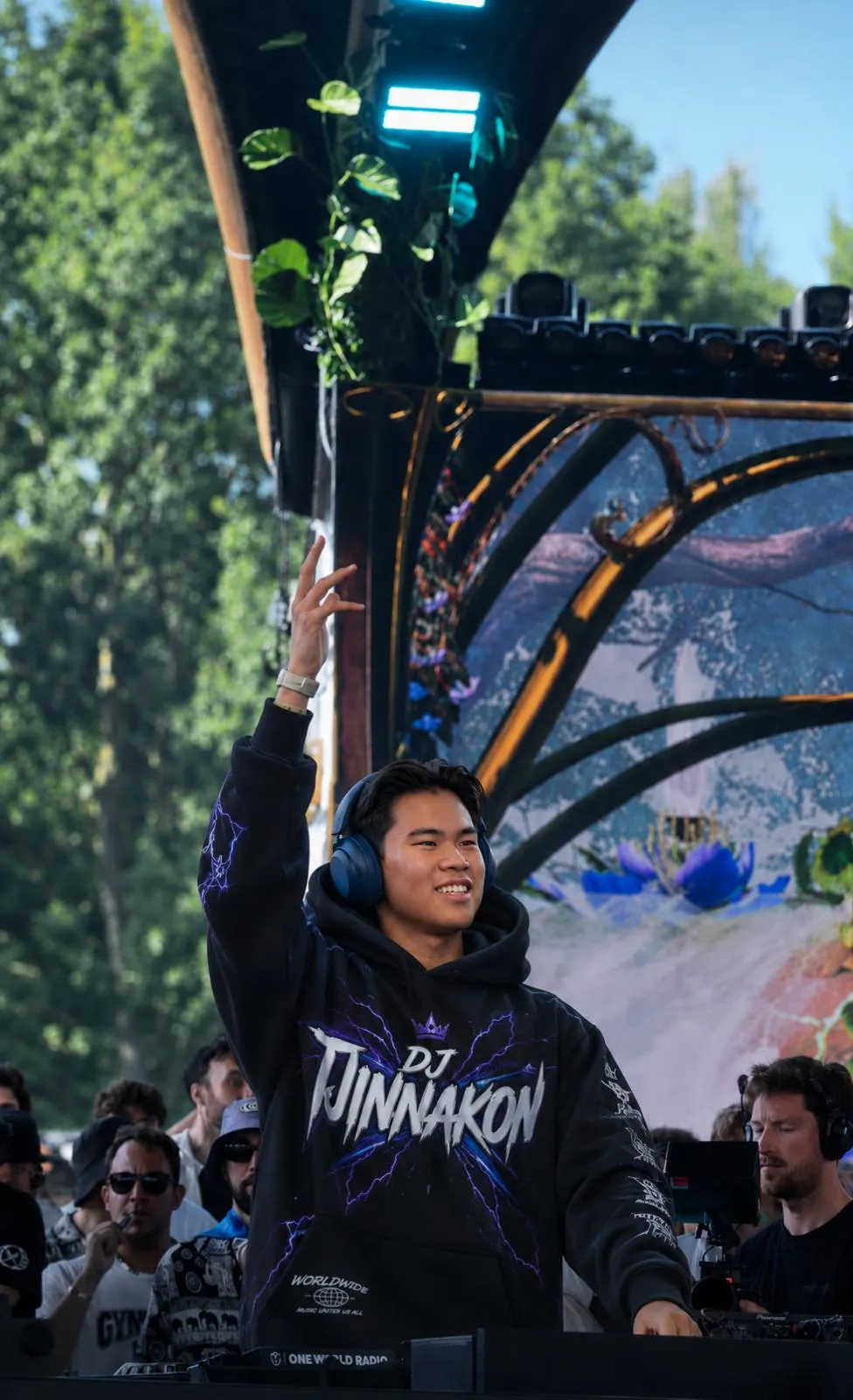 Tjinnakon steekt zijn hand omhoog tijdens een optreden op een festivalpodium, met publiek op de voorgrond.